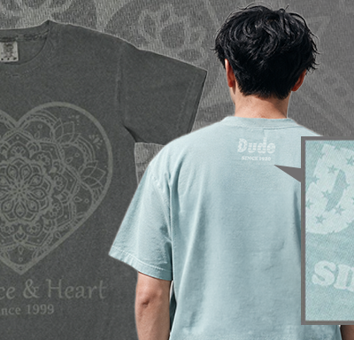 Back view of a person wearing a light blue T-shirt with a 'Dude' logo on the upper back; a dark gray T-shirt with a heart mandala and 'Essence & Heart Since 1999' design appears in the background.