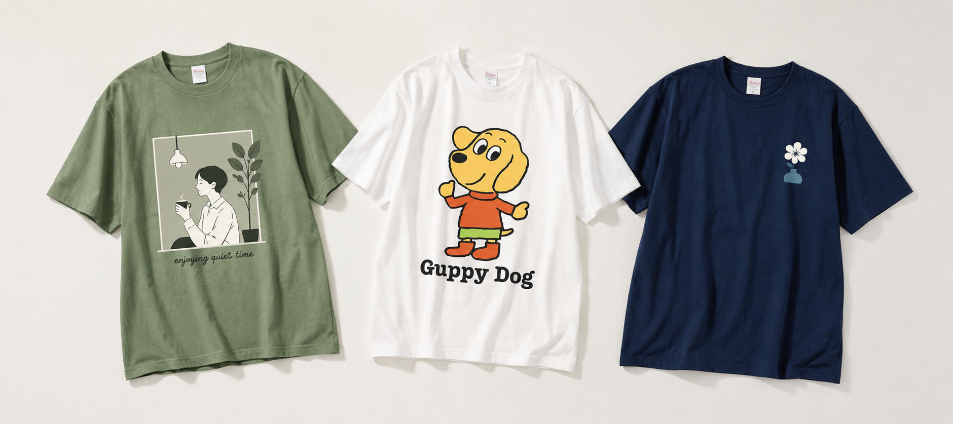 Three T-shirts laid flat: green tee with a framed illustration of a person sipping coffee beside a plant and 'enjoying quiet time' text, white tee with a Guppy Dog cartoon, and navy tee with a small flower in a pot graphic.